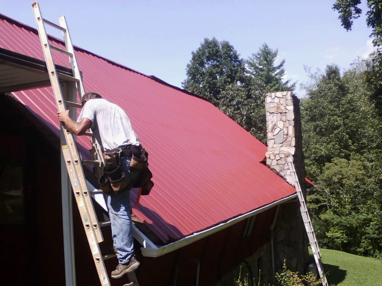 Roofer on a ladder beside a red metal roof and stone chimney — Soffit & Fascia Repair by Roofing Brothers in Freeville