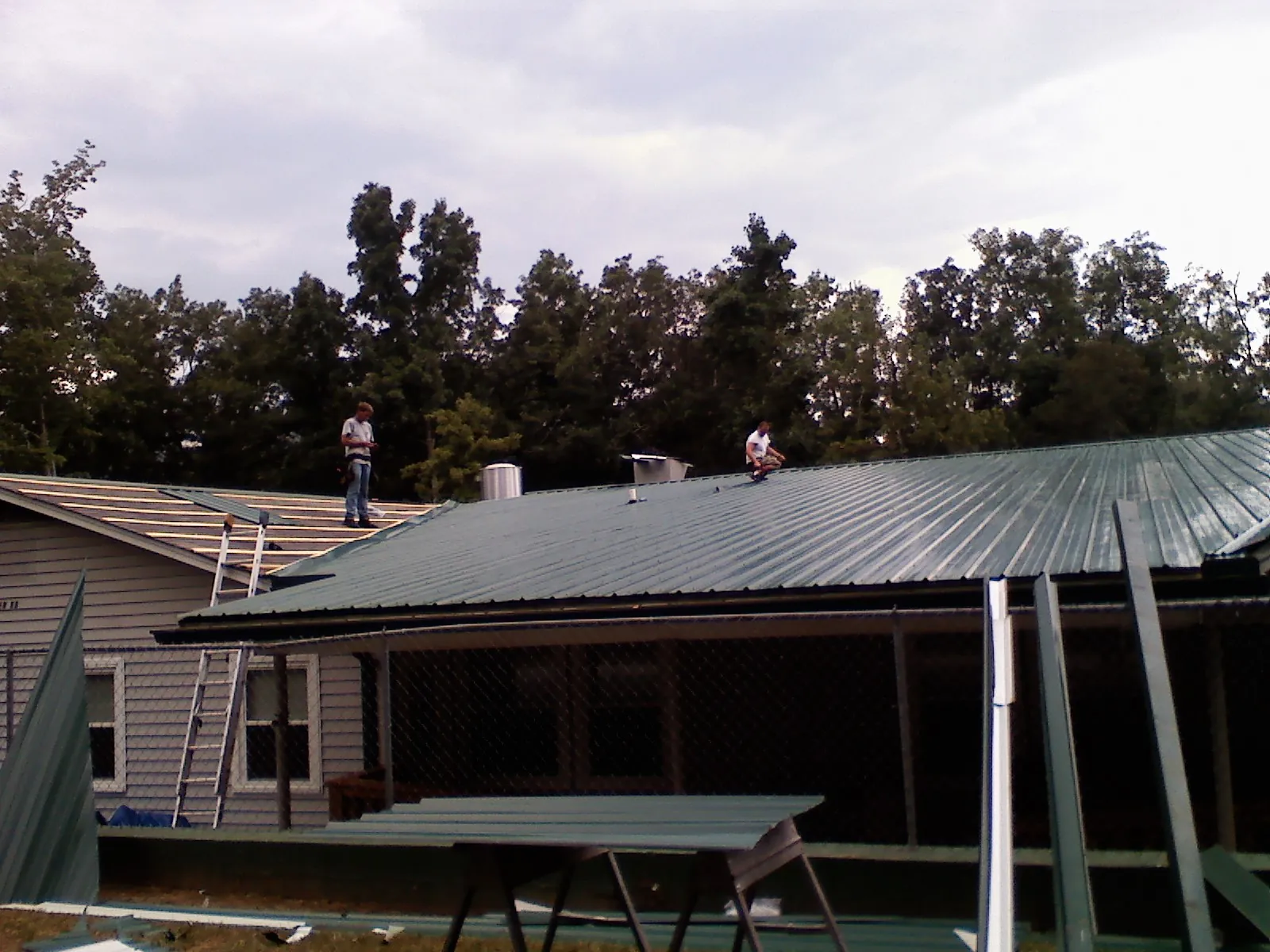 Two roofers installing green metal panels above a covered porch in Freeville